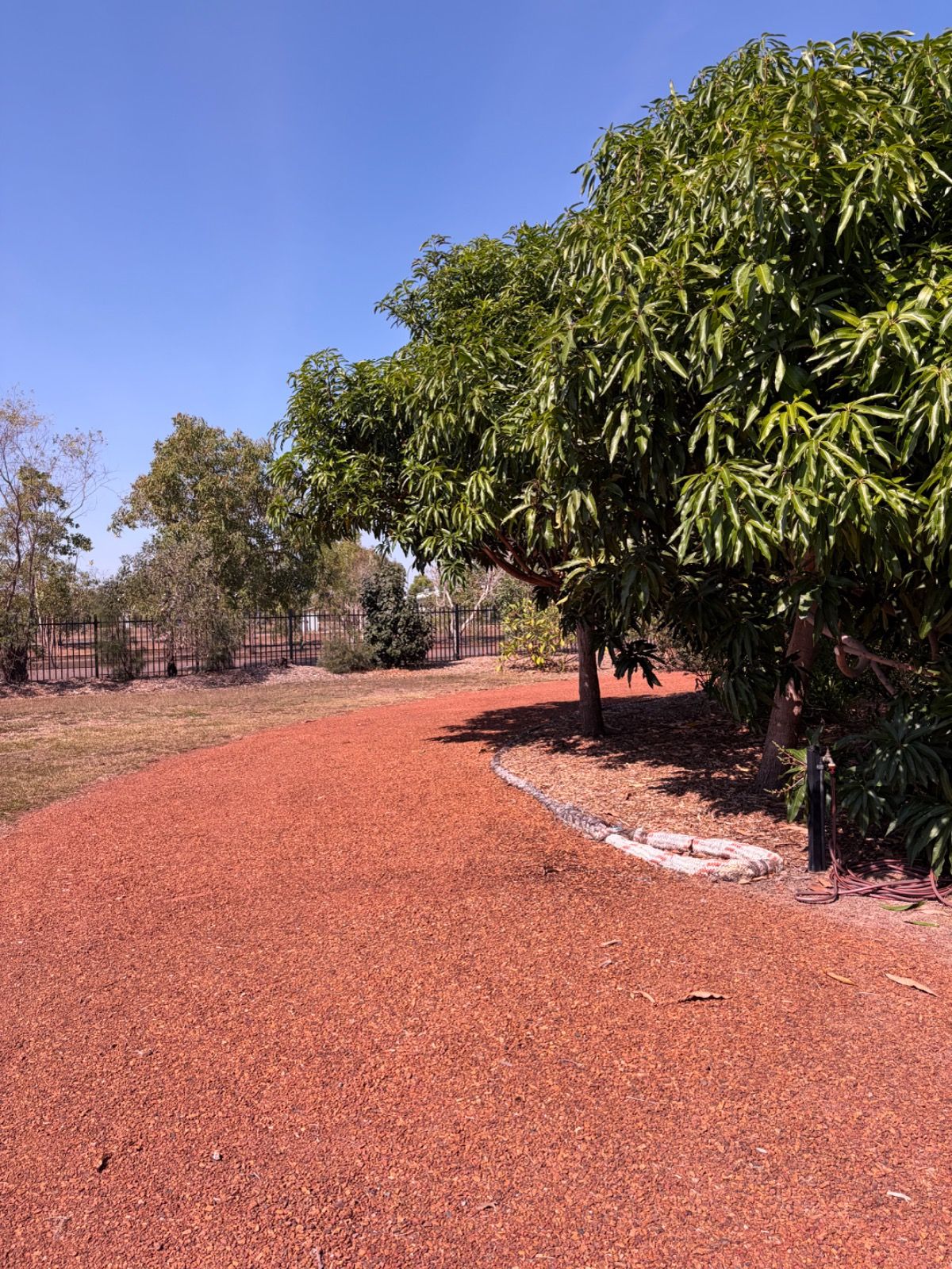 Curved red gravel pathway bordered by green leafy trees under a clear blue sky in a landscaped garden.