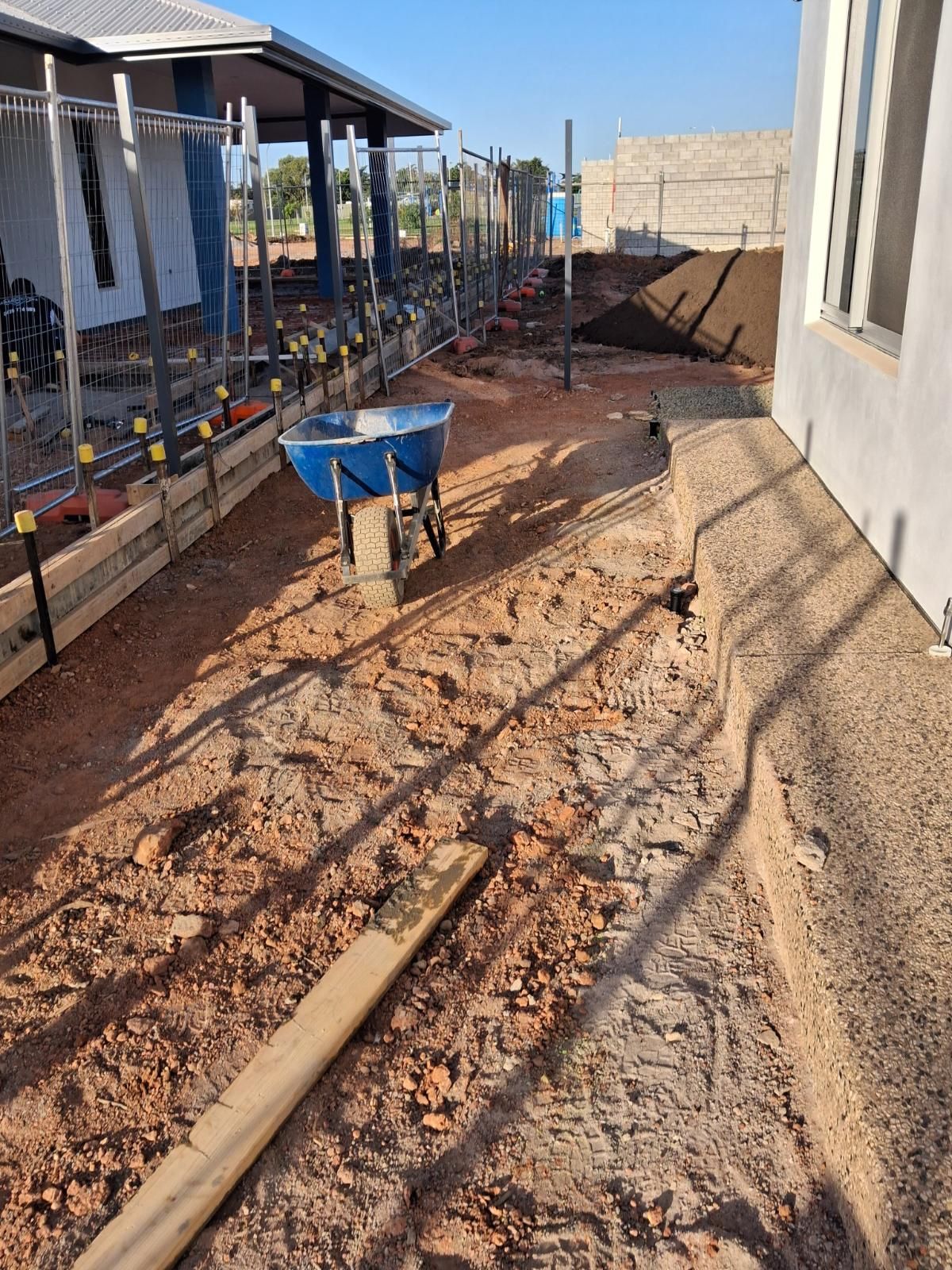 Cleared and levelled soil area with a blue wheelbarrow, wooden plank, and metal fencing along a house exterior in Marrara Gardens Northern Territory.