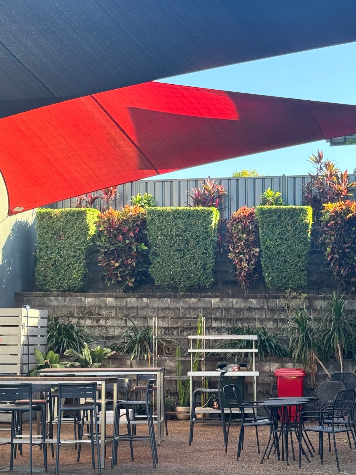 Outdoor seating area with metal tables and chairs under red and black shade sails, surrounded by trimmed hedges and colourful garden plants against a stone reta