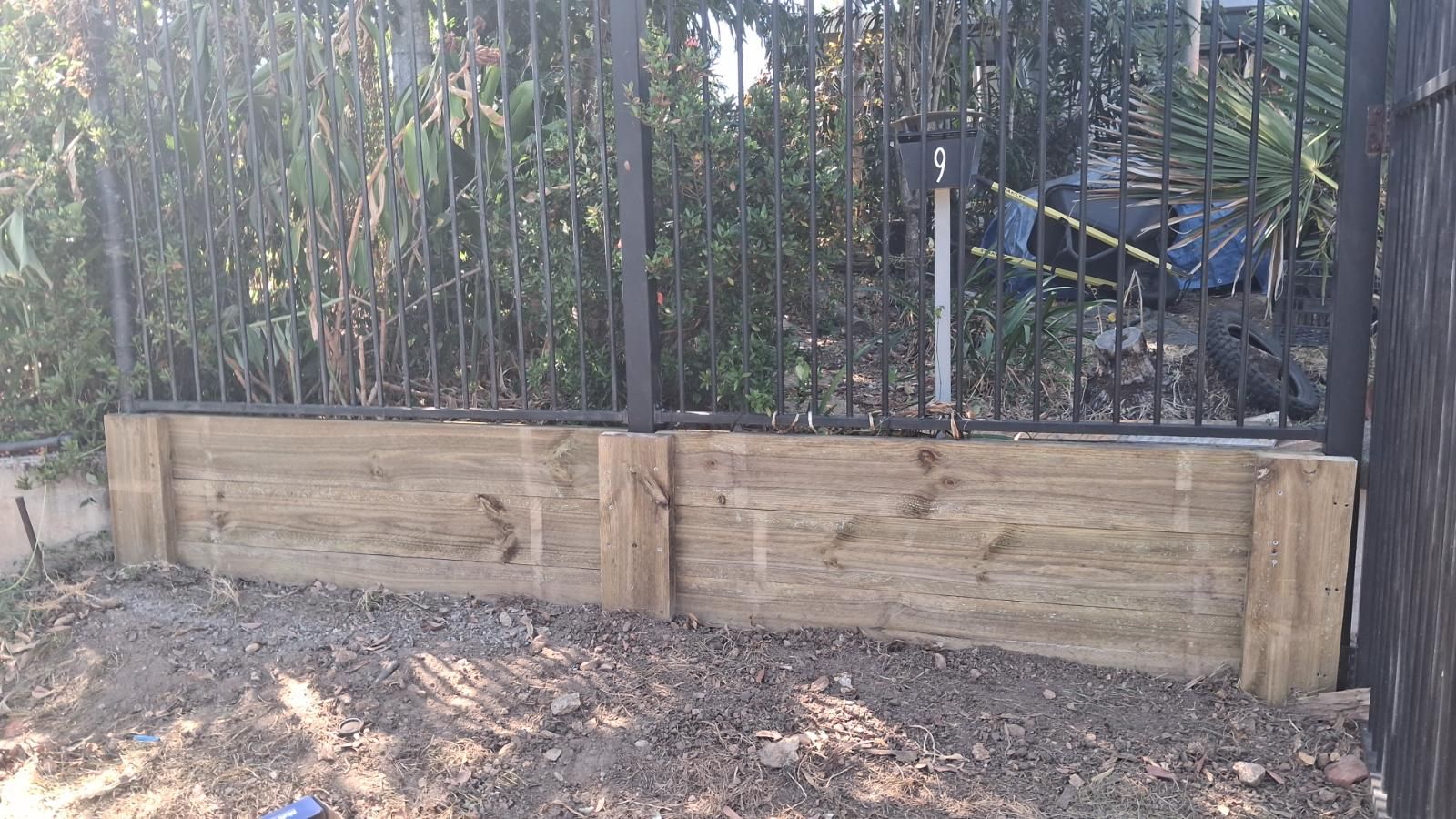 Freshly installed timber boundary panels fixed at the base of a black metal fence with plants behind and soil in front.