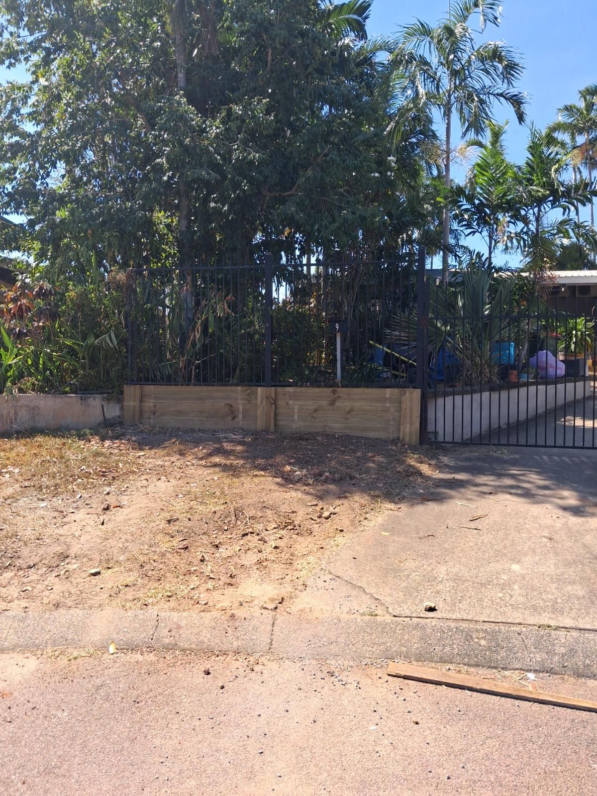Garden area with timber edging and black metal fence surrounded by trees and plants under clear blue sky