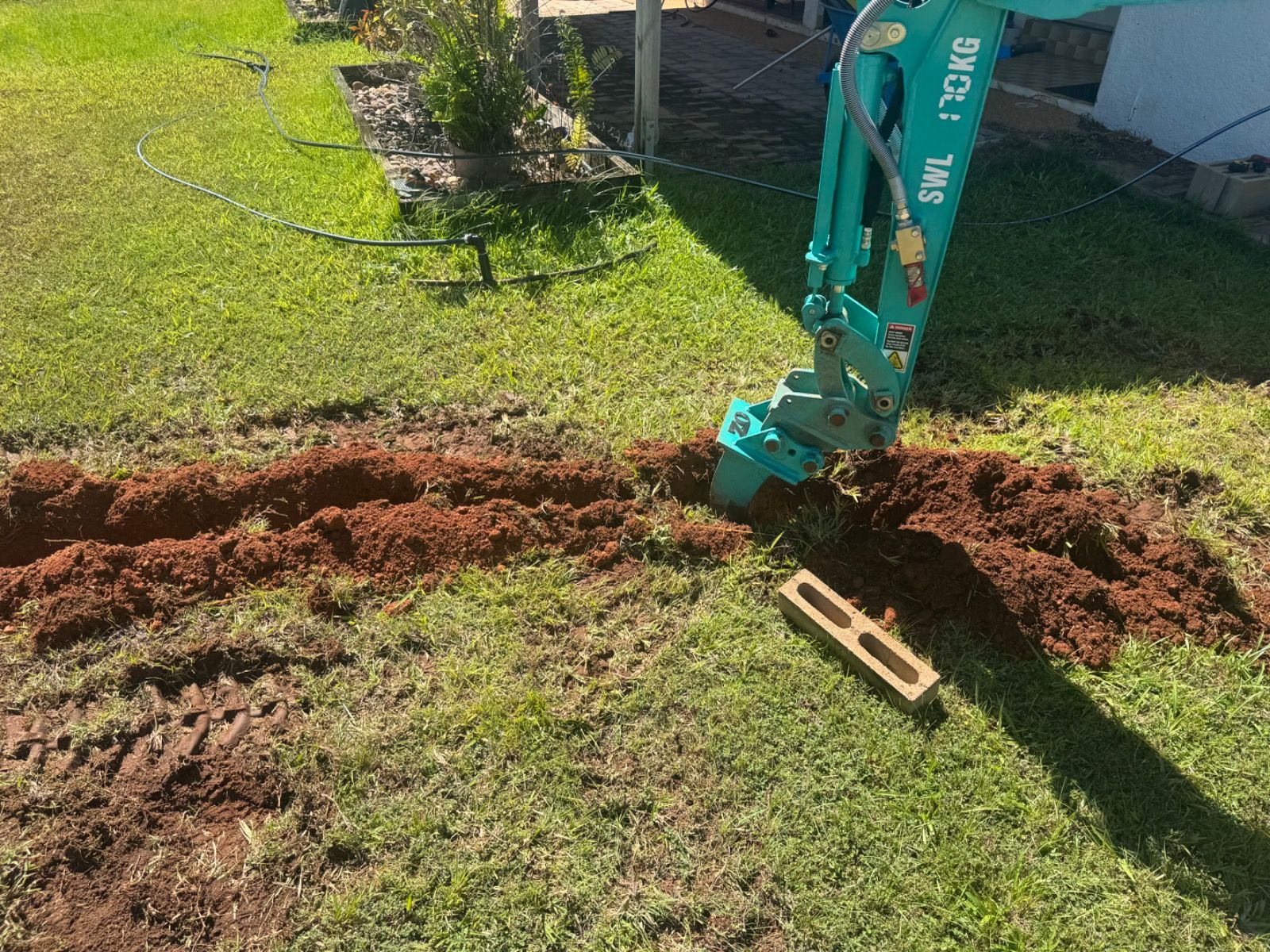 Mini excavator digging a narrow soil trench in green lawn garden in Leanyer Northern Territory