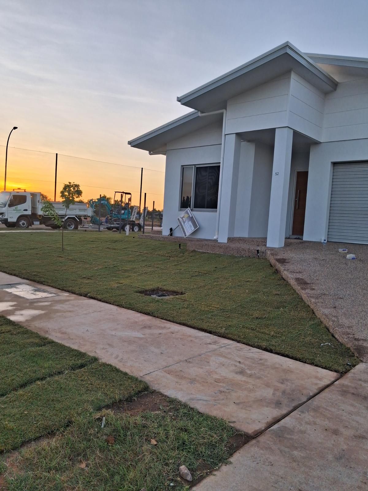 Front yard with freshly laid green turf and paved concrete path outside a modern white house during sunset