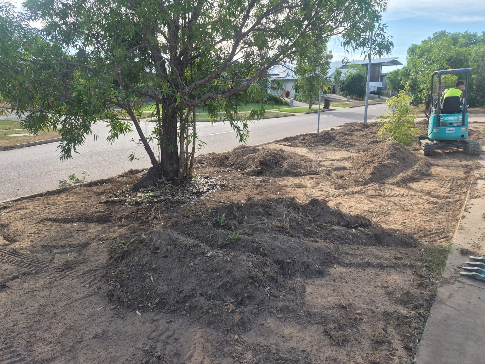 Garden beds cleared and fresh soil prepared around mature trees beside a road with a worker on a mini excavator in the background.